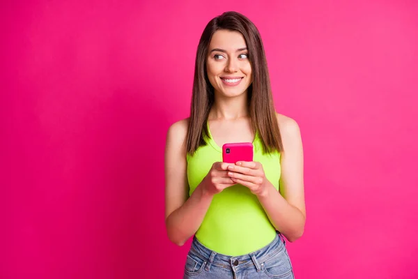Фото позитивної зацікавленої дівчини тримає мобільний телефон виглядає збоку порожній простір носіння зеленого сингла ізольований яскраво-рожевий кольоровий фон — стокове фото