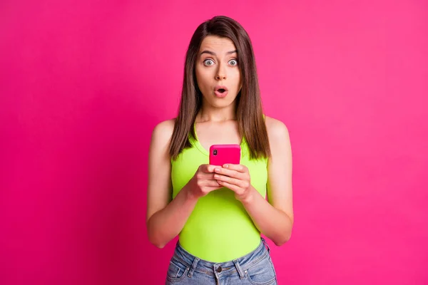 照片上兴奋的年轻女士拿着手机，瞪着眼张嘴，身穿绿色单色孤零零的亮粉色底色 — 图库照片