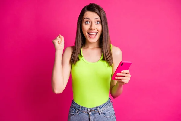 Photo de jeune femme étonnée tenir téléphone cellulaire bouche ouverte lever poing porter vert top isolé vibrant fond de couleur rose — Photo