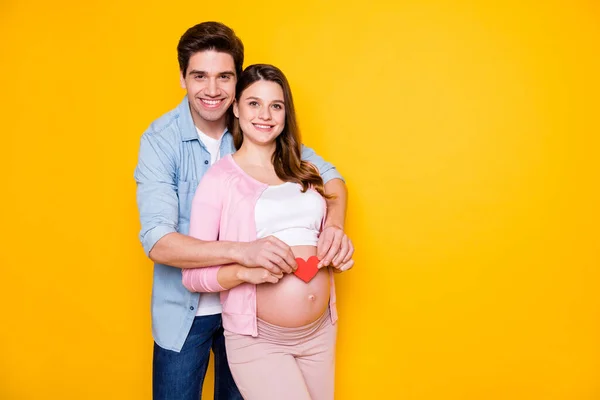 Foto do jovem casal stand abraço mulher grávida mostrar papel coração desgaste casual pano isolado no fundo amarelo — Fotografia de Stock