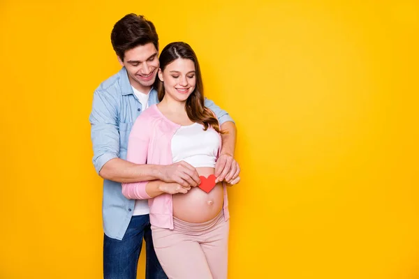 Foto de jovem casal stand abraço mulher grávida mostrar papel coração desgaste casual roupas isoladas no fundo de cor amarela — Fotografia de Stock
