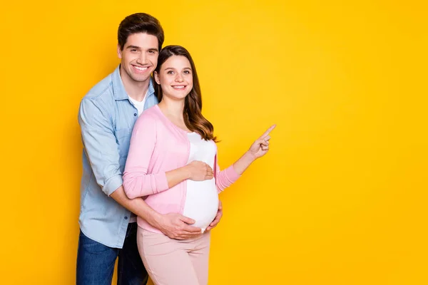 Foto de jovem casal stand abraço mulher grávida ponto vazio espaço desgaste casual roupas isoladas no fundo cor amarela — Fotografia de Stock