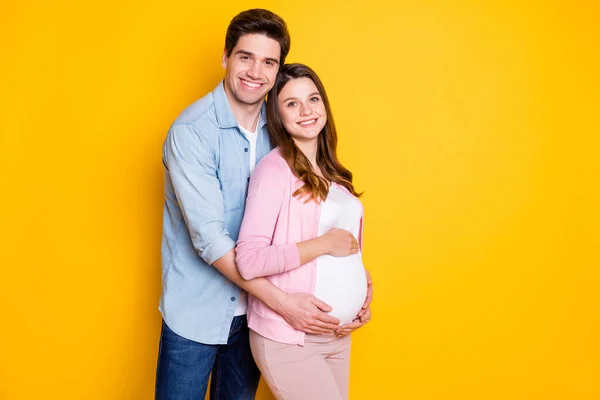 Foto de otimista bonito jovem casal stand abraço mulher grávida usar roupas casuais isolado no fundo de cor amarela — Fotografia de Stock