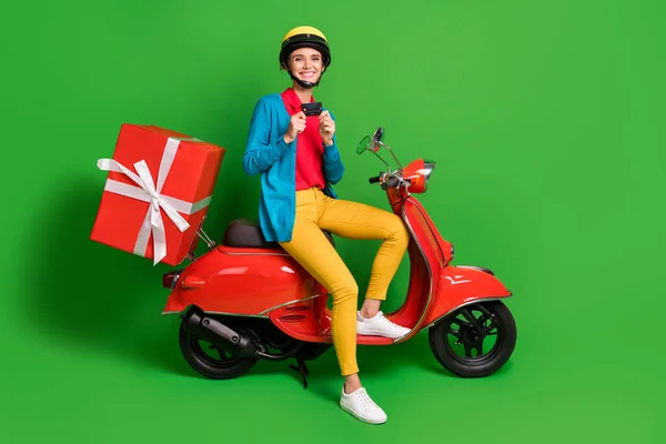 Pełny rozmiar zdjęcie młodej uśmiechniętej wesołej dziewczyny siedzieć czerwony motorower z dużą obecną posiadają kartę kredytową izolowane na zielonym tle kolor — Zdjęcie stockowe