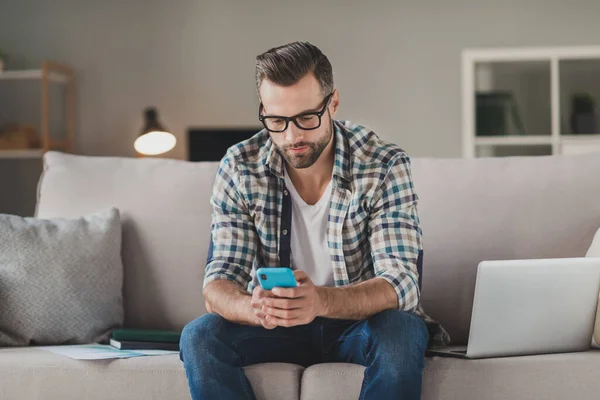 Фото молодої людини серйозно сидить диван додому дивіться читайте в інтернеті мобільний телефон носіння повсякденного вбрання фрілансер — стокове фото