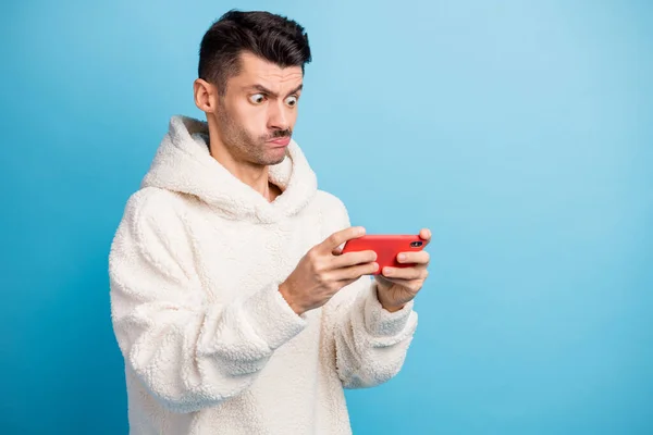 Retrato fotográfico de un hombre concentrado en muecas sosteniendo el teléfono en dos manos usando una sudadera con capucha de lana aislada sobre fondo de color azul pastel —  Fotos de Stock