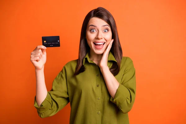 Fénykép őrült funky fiatal hölgy bemutató hitelkártya nyitott száj tenyér arc viselni zöld ing elszigetelt narancs színű háttér — Stock Fotó