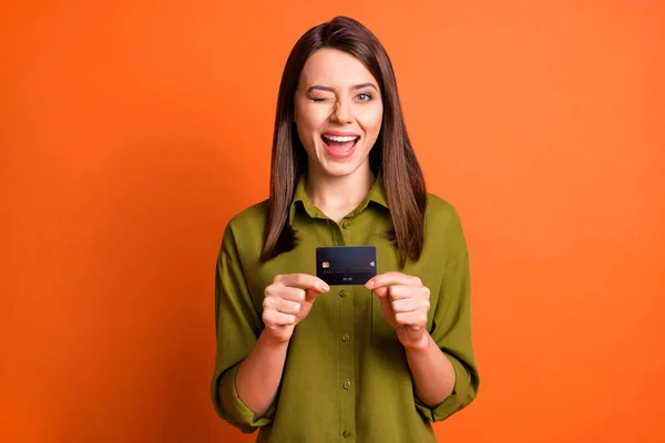 Foto de chica excitada coqueta presentando tarjeta de crédito cerrar guiño ojo desgaste camisa verde aislado color naranja fondo — Foto de Stock