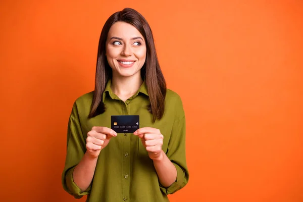 Foto de chica joven divertida mantenga tarjeta de débito mirada lado espacio vacío desgaste camisa verde aislado color naranja fondo — Foto de Stock