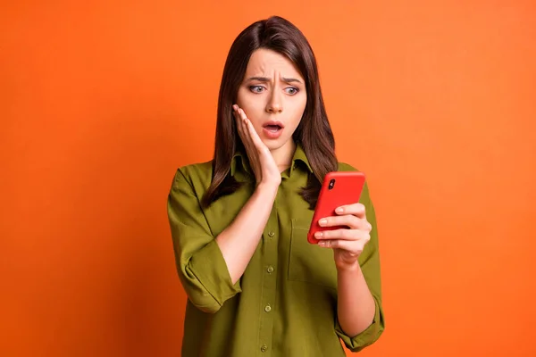 Photo of frightened young girl hold cellphone arm cheekbone look screen wear green shirt isolated orange color background — Stock Photo, Image