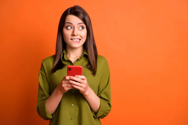Foto van schuldig jong meisje houden smartphone bijten lip kijken kant lege ruimte dragen groen shirt geïsoleerde oranje kleur achtergrond — Stockfoto