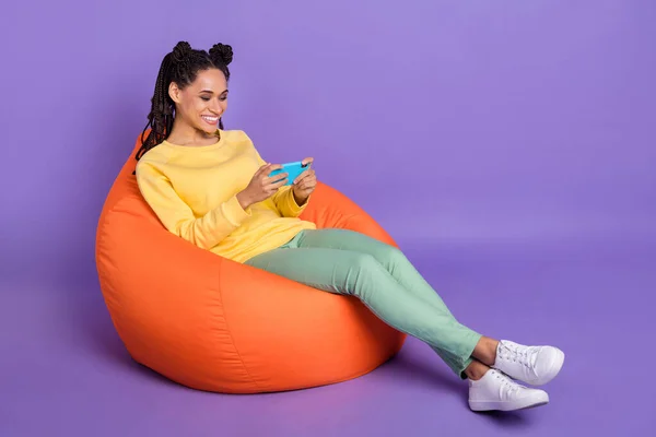 Full size photo of optimistic girl lying on pouf play telephone wear yellow shirt trousers sneakers isolated on lilac color background — Stock Photo, Image