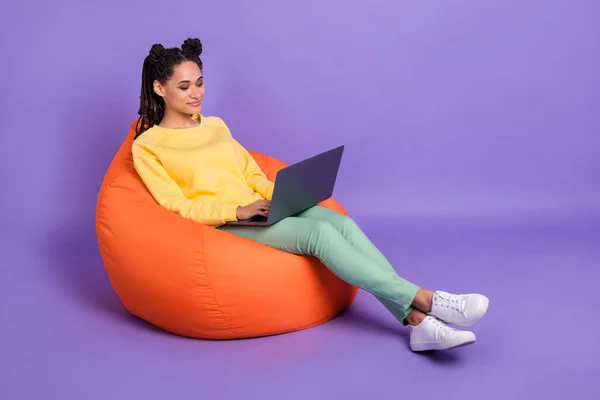 Foto de tamaño completo de chica optimista acostado en el aspecto del puf portátil usar pantalones de camisa amarilla zapatillas aisladas en color lila fondo —  Fotos de Stock