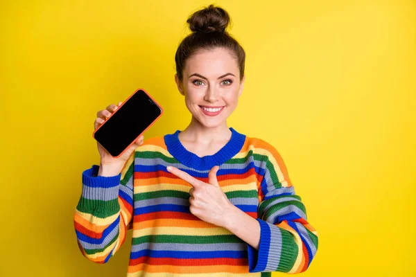 Photo of pretty brown bun haired lady point phone screen copyspace wear rainbow sweater isolated on yellow color background