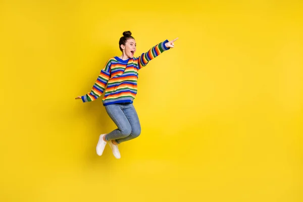 Foto de cuerpo completo de moño marrón chica punta dedo mirada copyspace saltar usar pantalones vaqueros suéter de arco iris aislado sobre fondo de color amarillo — Foto de Stock