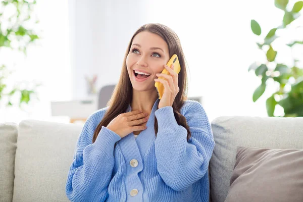 Портрет довольно веселой девушки, сидящей на диване и разговаривающей по телефону, обсуждающей смешные хорошие новости в светлой квартире — стоковое фото