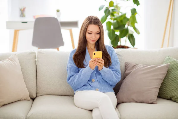 Retrato de atractiva chica de pelo castaño enfocado sentado en diván usando aplicación de chat dispositivo en casa plana de luz apartamento interior — Foto de Stock