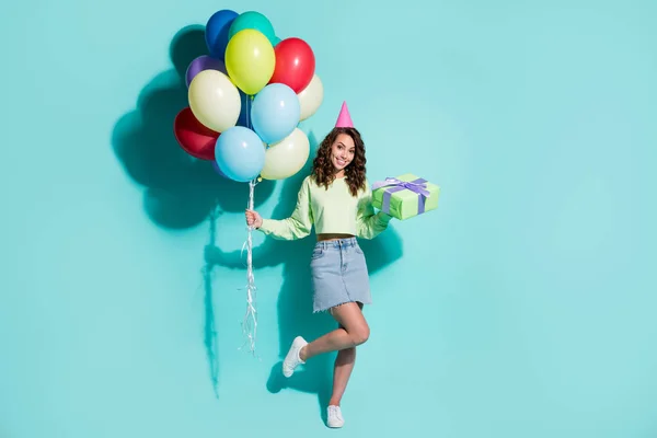 Foto von charmanten Mädchen halten Geschenk Haufen Luftballons tragen Kegelmütze grünen Pullover Jeans Mini Rock Turnschuhe isoliert blaue Farbe Hintergrund — Stockfoto