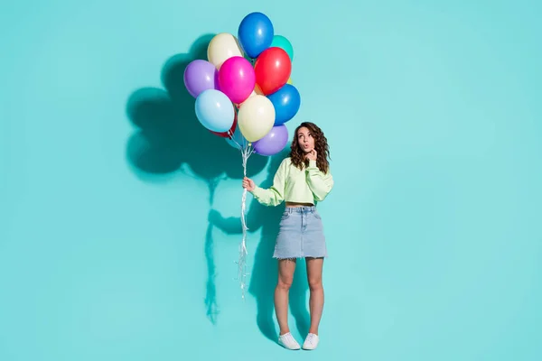 Foto da menina segurar muitos balões dedo bochecha olhar espaço vazio desgaste camisola verde mini jeans saia tênis isolado azul cor fundo — Fotografia de Stock