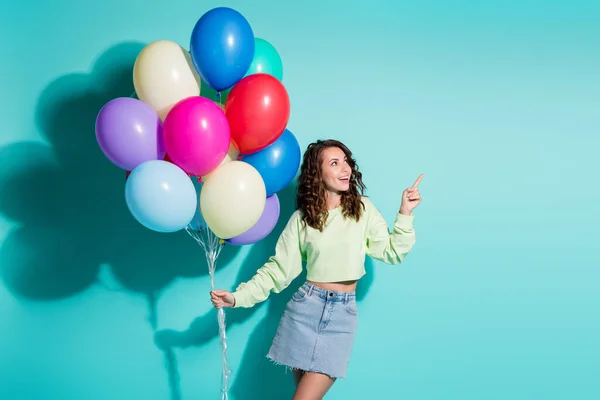 Foto di cute ragazza tenere molti palloncini indicano dito sguardo vuoto spazio usura verde maglione mini jeans gonna isolato colore blu sfondo — Foto Stock