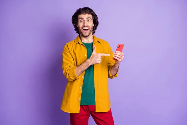 Zdjęcie młody podekscytowany szczęśliwy uśmiechnięty dobry nastrój mężczyzna facet wskazując palcem na smartfonie odizolowany na fioletowym tle koloru — Zdjęcie stockowe