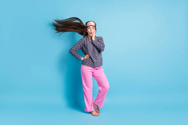 Full length photo of cute teenager girl air blow long hairdo barefoot palm face shock weather change sleep wear mask dotted shirt pink pants pajamas sleepwear isolated blue color background