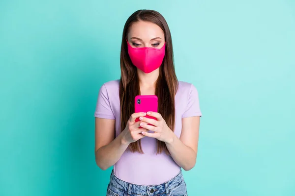 Foto retrato de menina vestindo rosa máscara de tecido protetor mensagens de texto no smartphone isolado no fundo de cor teal brilhante — Fotografia de Stock