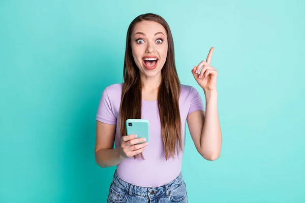 Фотопортрет изумленной девушки, держащей смартфон, смотрящей вверх пальцем, изолированным на ярком бирюзовом фоне — стоковое фото