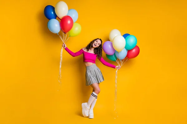 Foto de cuerpo entero del estudiante bastante femenino sosteniendo globos de aire en ambas manos riendo alegremente usando falda a cuadros aislada sobre fondo de color amarillo brillante — Foto de Stock