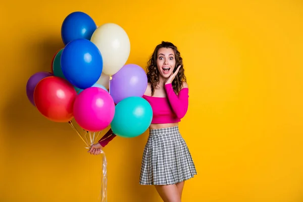 Foto de retrato de chica sorprendida sosteniendo globos de aire coloridos manteniendo la mano limpia cara con la boca abierta sonriendo usando falda a cuadros aislada sobre fondo de color amarillo brillante — Foto de Stock