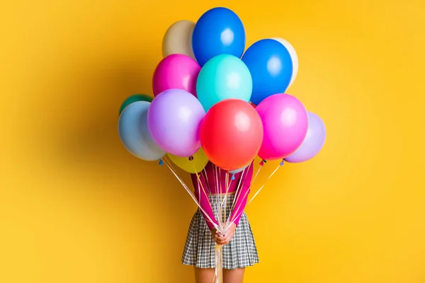 Foto retrato de menina engraçada fazendo surpresa escondendo rosto mantendo balões coloridos vestindo roupas casuais isoladas no fundo de cor amarela — Fotografia de Stock