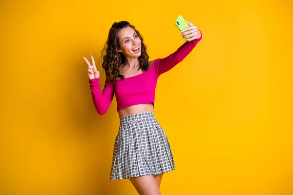 Photo of lovely pretty lady faliste hairdo trzymać telefon make selfie show v-sign toothy smile nosić różowy top rozbierane ramiona mini spódnica odizolowany żółty kolor tło — Zdjęcie stockowe