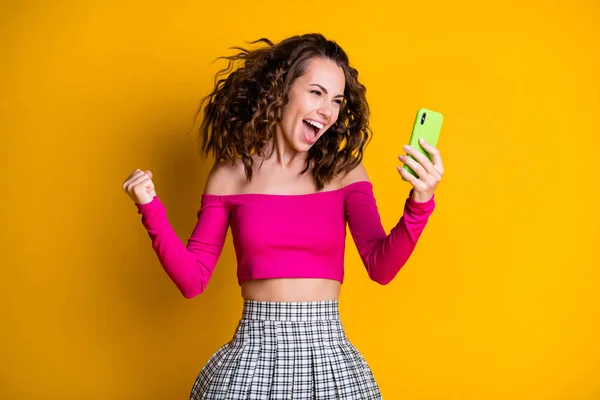 Foto de linda dama mantener teléfono rizado peinado loco boca abierta triunfo emocionada lotería ganar desgaste rosa superior sin ropa hombros mini falda aislado color amarillo fondo — Foto de Stock