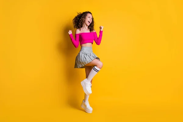 Φωτογραφία πορτρέτο του κοριτσιού γιορτάζει τη νίκη στέκεται στο ένα πόδι γροθιές επάνω φορώντας casual ροζ καλλιέργεια-top καρό φούστα μακρύ κάλτσες λευκά αθλητικά παπούτσια απομονώνονται σε ζωντανό κίτρινο χρώμα φόντο — Φωτογραφία Αρχείου