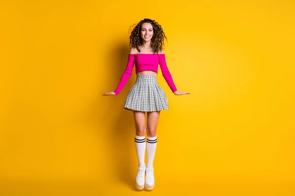 Foto ritratto di felice ragazza che salta le mani ai lati indossando rosa crop-top gonna a quadri calze lunghe scarpe da ginnastica bianche isolate su sfondo giallo vivido — Foto Stock