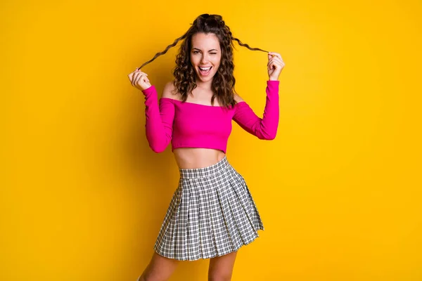Foto portrét funky dívka drží dva vlasy zámky s rukama mrkání se směje na sobě růžové plodiny-top kostkované mini sukně izolované na jasně žluté barevné pozadí — Stock fotografie