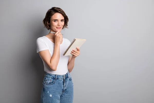 Foto de joven atractiva mujer positiva alegre sostener diario de cuaderno escribir mano tacto mentón aislado sobre fondo de color gris —  Fotos de Stock