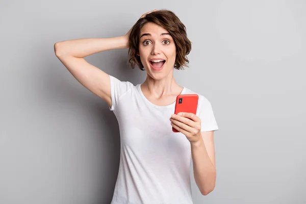 Foto van aantrekkelijke vrolijke jonge meisje houden mobiele telefoon chat blij opgewonden verrast hand touch hoofd geïsoleerd over grijze kleur achtergrond — Stockfoto