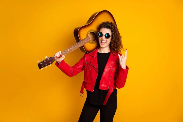 Retrato de atraente alegre alegre alegre menina de cabelos ondulados carregando guitarra mostrando sinal de chifre isolado no fundo de cor amarelo brilhante — Fotografia de Stock