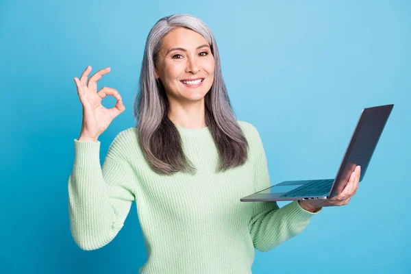 Photo de positive vieille dame tenir ordinateur portable montrer ok signe porter pull pull pull vert isolé sur fond de couleur bleue — Photo