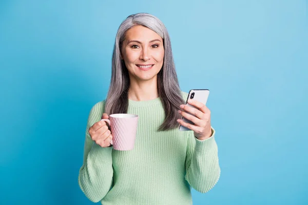 Photo of retired old woman arm hold cup smartphone look camera wear green sweater isolated blue color background — Stock Photo, Image