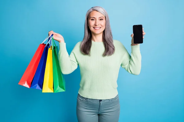 Retrato de atraente mulher de cabelos grisalhos alegre transportar pacotes demonstrar telefone desgaste casual isolado no fundo de cor azul — Fotografia de Stock