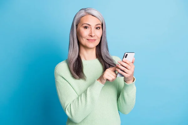 Porträt von attraktiven zufriedenen älteren Frau halten Handy-Eingabe gekleidet grünen Pullover isoliert auf blauem Hintergrund — Stockfoto
