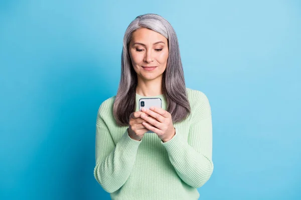 Elinde telefon tutan sevimli yaşlı bir kadının fotoğrafı mavi arka planda izole edilmiş yeşil kazak giymiş. — Stok fotoğraf
