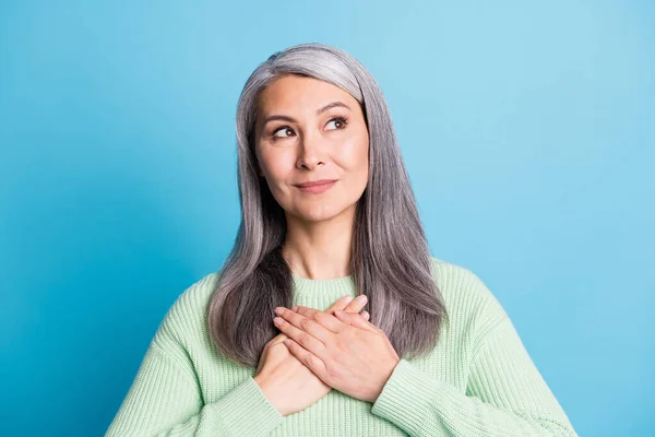 Foto von dankbaren verträumten reifen Dame suchen leeren Raum halten gekreuzte Handflächen auf Herz tragen grünen Pullover isoliert auf blauem Hintergrund — Stockfoto