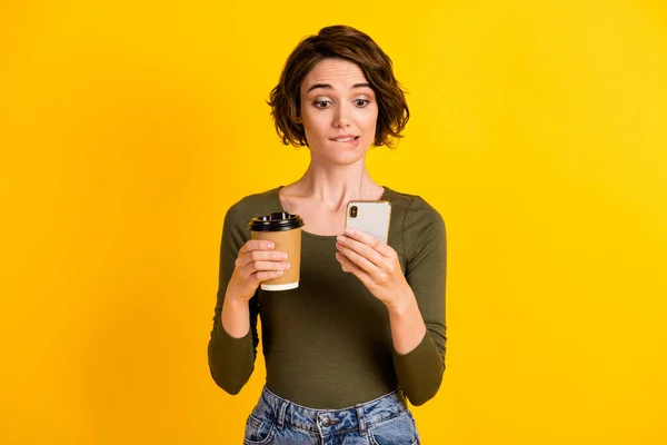 Foto de linda chica mantenga teléfono taza bebida mordida labio pantalla desgaste camisa verde aislado vívido color amarillo fondo — Foto de Stock