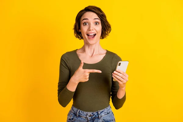 Foto de niña divertida sostener teléfono dedo directo boca abierta sorprendido usar camisa verde aislado vívido color amarillo fondo — Foto de Stock