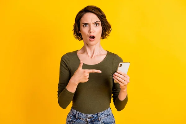 Photo of young lady hold smartphone indicate finger open mouth staring mad wear green shirt isolated bright yellow color background