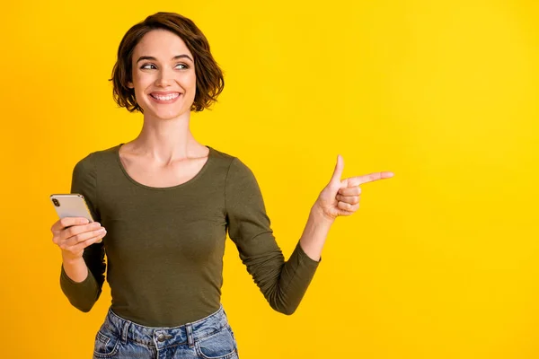 Foto de teléfono inteligente de retención de chica bonita indican mirada de dedo espacio vacío desgaste camisa verde aislado vibrante color amarillo fondo — Foto de Stock