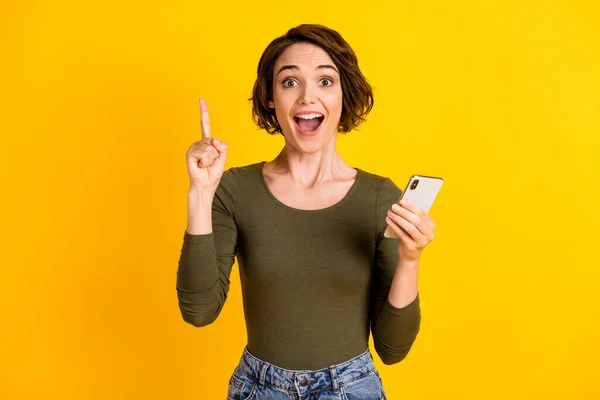 Foto di giovane ragazza tenere telefono alzare indice bocca aperta indossare camicia verde isolato vibrante colore giallo sfondo — Foto Stock